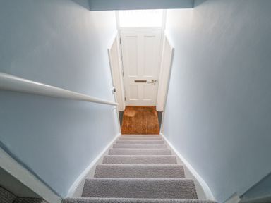 A staircase with carpet leading down to a white front door with a doormat at The Corner Cottage in Aldeburgh