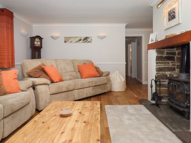 A living room with beige sofas and orange pillows a wooden coffee table a stone fireplace and a grandfather clock at Hole Park in Holsworthy