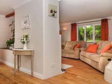 A living room with two beige sofas and orange cushions near a window with orange curtains and a small table with flowers against a white wall at Hole Park in Holsworthy