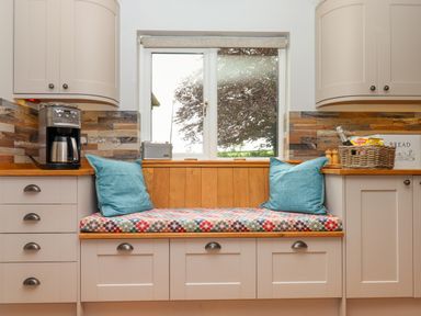 A kitchen window seat with blue cushions and a patterned cushion between kitchen cabinets with countertop appliances and a basket at Hole Park in Holsworthy