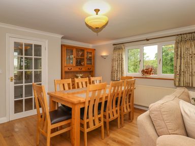 A dining room with a wooden table and chairs a wooden cabinet beige sofa and a window with curtains at Hole Park in Holsworthy