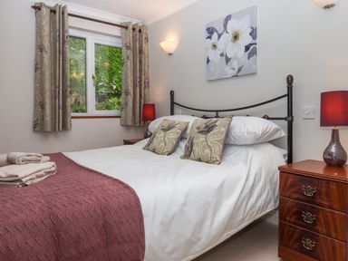 A bedroom with a double bed with floral pillows a maroon blanket bedside tables with lamps and a window with floral curtains at Hole Park Holsworthy