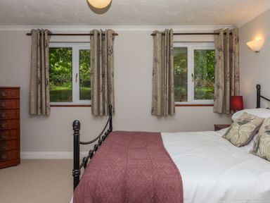 A bedroom with a bed covered with pillows and a blanket two windows with curtains a wooden chest of drawers and a bedside table with a lamp at Hole Park in Holsworthy