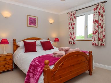 A bedroom with a wooden bed with white bedding and red pillows next to a window with floral curtains at Hole Park in Holsworthy