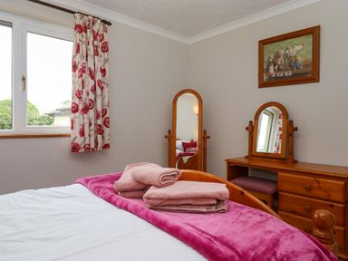 A bedroom with a bed with folded towels and a pink blanket a wooden dressing table with mirrors a striped stool floral curtains on a window and a framed picture on the wall at Hole Park in Holsworthy