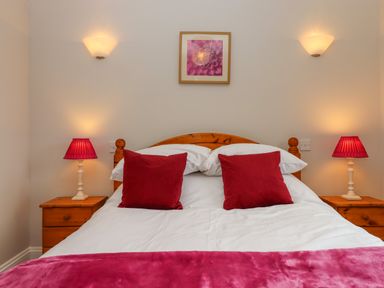 A bedroom with a wooden bed with white bedding and red pillows beside wooden nightstands with red lamps and a pink flower picture on the wall at Hole Park in Holsworthy