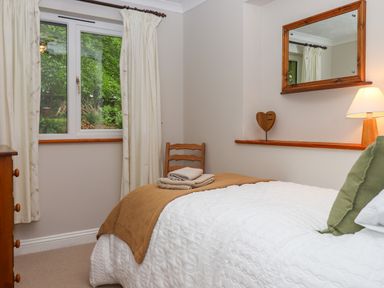 A bedroom with a bed covered in white and brown blankets green pillow a wooden chair folded towels a window with curtains a wooden dresser a mirror and a lamp on a shelf at Hole Park in Holsworthy