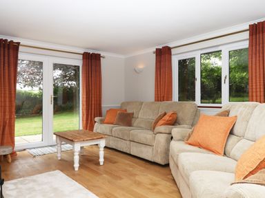 A living room with two beige sofas orange and brown cushions a wooden coffee table and glass doors with orange curtains at Hole Park in Holsworthy