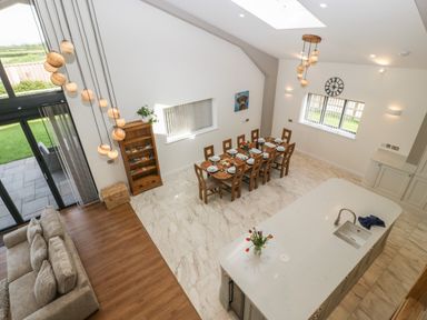 A dining area with a table set for eight people next to a kitchen island with a sink and a vase of flowers in an open plan room with a sofa and large windows at Hidden Well in Aust
