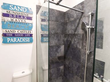 A bathroom with a shower area with dark grey tiles and a wall decoration with beach words at Kingfisher Cottage in Ottinge near Lyminge