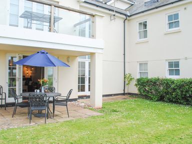 A patio area with a round table and chairs with a blue umbrella in a grassy courtyard at 5 Thurlestone Beach in Thurlestone
