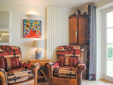 A sitting area with patterned armchairs a wooden side table with a lamp and bookshelf a wooden corner cabinet and a colorful octopus painting at 5 Thurlestone Beach in Thurlestone