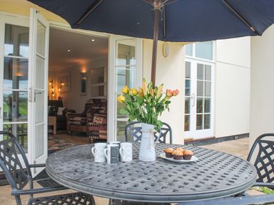 An outdoor patio table with a vase of flowers four mugs and a plate of muffins under a large umbrella next to open glass doors at 5 Thurlestone Beach in Thurlestone