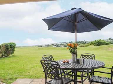 An outdoor patio with a round table and chairs under a blue umbrella with flowers and muffins on the table at 5 Thurlestone Beach in Thurlestone
