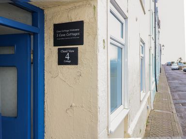 A cream colored cottage wall with a blue door and signs for Cove Cottage Hideaway and Chesil View 4 Cottage on a street with parked cars at Cove Cottage Hideaway in Chiswell On Portland