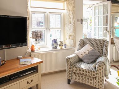 A living room with a patterned armchair by a window with vases and a small table with a television at Cove Cottage Hideaway in Chiswell On Portland