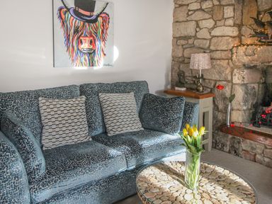 A sitting area with a patterned blue sofa and cushions a round stone top coffee table with flowers a stone fireplace and a side table with a lamp at Cove Cottage Hideaway Chiswell On Portland
