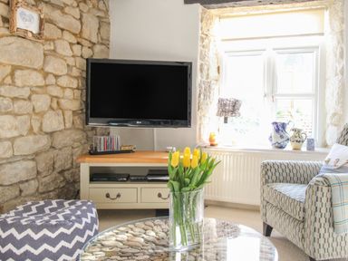 A living room with a stone wall a TV on a cabinet a vase with yellow tulips a patterned armchair and a window at Cove Cottage Hideaway in Chiswell On Portland
