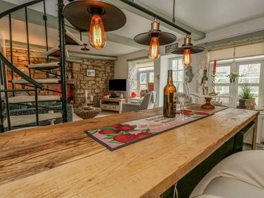 A living room with a wooden table, spiral staircase, stone wall, armchair, and windows at Cove Cottage Hideaway in Chiswell On Portland