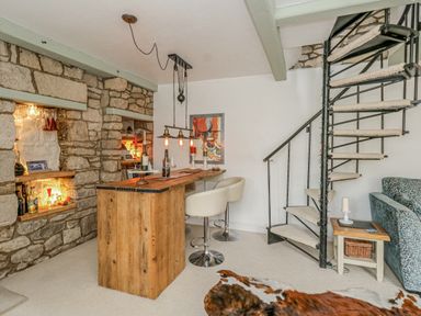 A room with a stone wall, wooden bar table with stools, spiral staircase, and a couch at Cove Cottage Hideaway in Chiswell On Portland
