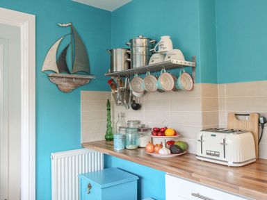 A kitchen corner with a wooden countertop cups hanging on a metal rack pots on a shelf a toaster and bowls of vegetables at Cove Cottage Hideaway in Chiswell On Portland