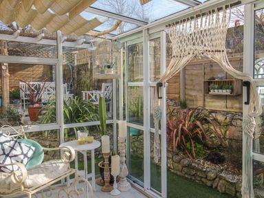 A sunroom with plants candles and a chair with cushions overlooking a garden at Cove Cottage Hideaway in Chiswell On Portland