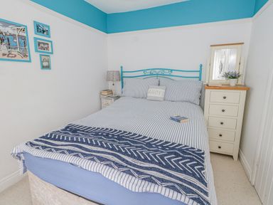 A bedroom with a bed with striped bedding a bedside table with a lamp and books a chest of drawers with a mirror and a flower pot and pictures on the wall at Cove Cottage Hideaway in Chiswell On Portland