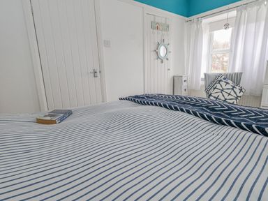 A bedroom with a striped bedspread a book on the bed a chair with pillows a window and curtains at Cove Cottage Hideaway in Chiswell On Portland