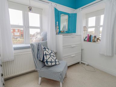 A corner room with two windows featuring white curtains a striped chair with cushions a white chest of drawers and a blue shuttered mirror at Cove Cottage Hideaway Chiswell On Portland