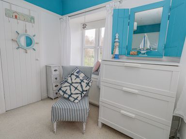 A room with a striped chair, a small drawer unit, a window with white curtains and a white dresser with a blue mirror at Cove Cottage Hideaway Chiswell On Portland