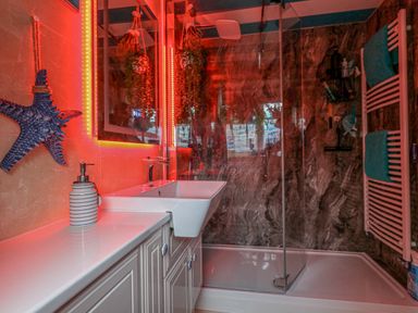A bathroom with a sink and countertop a glass-enclosed shower with dark marble walls and a white towel rack with blue towels at Cove Cottage Hideaway in Chiswell On Portland