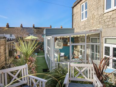 A garden seating area with white chairs next to a glass conservatory attached to a stone house at Cove Cottage Hideaway in Chiswell On Portland
