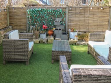 An outdoor seating area with wicker chairs and a table on artificial grass with a water fountain and plants at Cove Cottage Hideaway in Chiswell On Portland