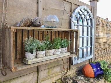 A wooden hanging shelf with small potted plants and pinecones beside a window and a large pottery jug on a stone surface at Cove Cottage Hideaway in Chiswell On Portland