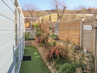 A narrow garden path with artificial grass beside a house and stone flower beds with plants at Cove Cottage Hideaway in Chiswell On Portland