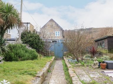 A garden with a stone pathway leading to a blue wooden gate and a stone house in the background at Cove Cottage Hideaway in Chiswell On Portland