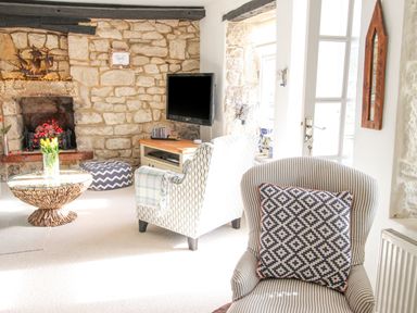A living room with a stone fireplace armchairs a round wooden coffee table and a wall-mounted TV at Cove Cottage Hideaway in Chiswell On Portland