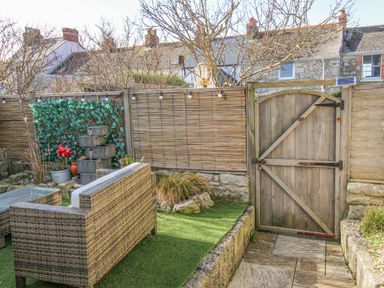 An outdoor garden area with wicker seating a wooden gate stone flooring and a water feature at Cove Cottage Hideaway in Chiswell On Portland
