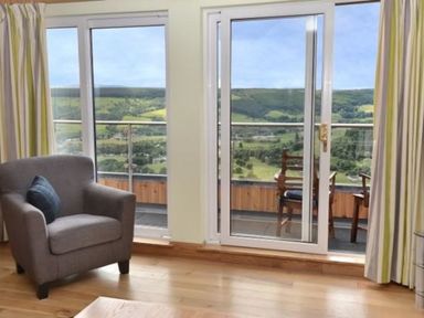 A room with a gray armchair and a balcony with outdoor chairs overlooking green hills at Glen View Lodge in Drumnadrochit Loch Ness