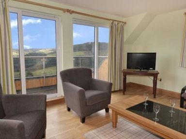 A living room with two armchairs a coffee table with glasses a TV on a stand and large windows overlooking a hillside at Glen View Lodge in Drumnadrochit Loch Ness