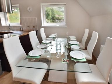 A dining area with a glass table set with plates and glasses and white chairs at Glen View Lodge in Drumnadrochit Loch Ness