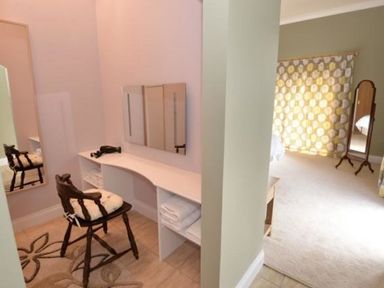 A corner dressing area with a wooden chair and a vanity mirror next to a bedroom with a standing mirror and patterned curtains at Glen View Lodge in Drumnadrochit Loch Ness