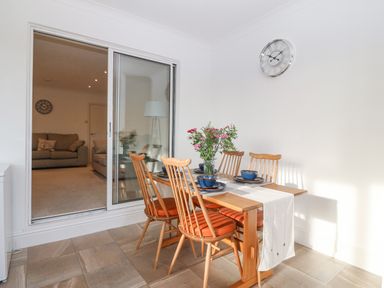 A dining area with wooden table and chairs set with blue bowls next to a sliding glass door leading to a living room with sofas at Riverview Apartment in Cemaes Bay