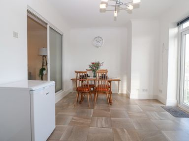 A dining area with a wooden table and four chairs set with plates and a flower vase in a room with tiled floor and a white refrigerator at Riverview Apartment in Cemaes Bay