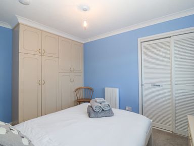 A bedroom with blue walls a bed with white sheets and folded towels beige wardrobes a wooden chair and white closet doors at Riverview Apartment in Cemaes Bay