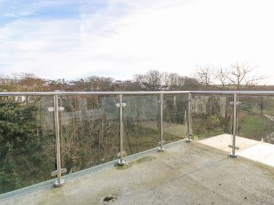 A balcony with glass and metal railing overlooking trees and distant houses at Riverview Apartment in Cemaes Bay