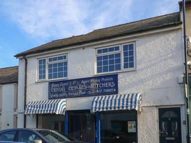 A two story white building with two blue and white striped awnings and a blue sign reading butchers with a car parked in front at Riverview Apartment in Cemaes Bay