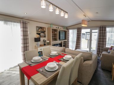 A dining table set for four with white dishes and a red runner in a living room with beige sofas and a TV unit at Ladybird Lodge South Lakes Leisure Park near Carnforth