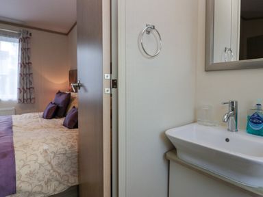 A bathroom sink with a mirror and soap dispenser next to a partially open door showing a bedroom with a bed and cushions at Ladybird Lodge South Lakes Leisure Park near Carnforth