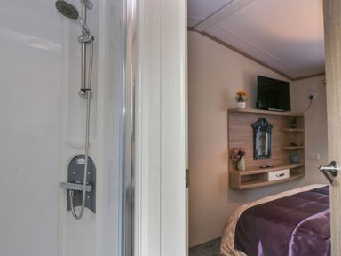 A shower area next to a bedroom with a wall-mounted television and a wooden shelf unit at Ladybird Lodge in South Lakes Leisure Park near Carnforth